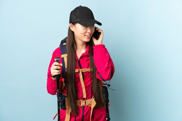 Niña china con mochila y bastones de trekking sobre azul aislado manteniendo una conversación con el teléfono móvil con alguien