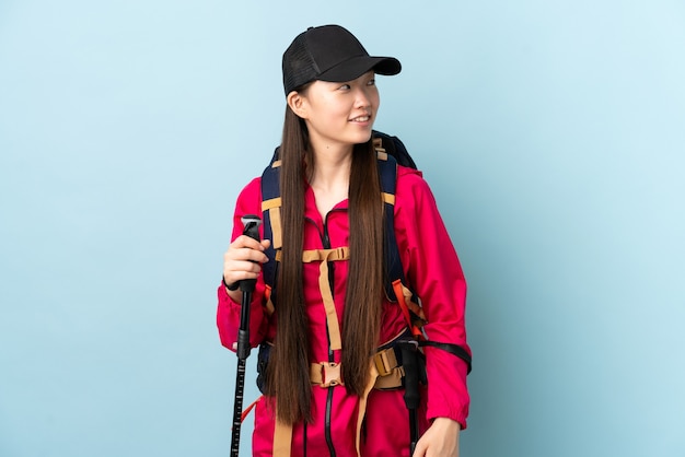 Niña china con mochila y bastones de trekking sobre azul aislado pensando en una idea mientras mira hacia arriba