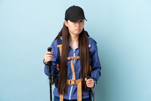 Niña china con mochila y bastones de trekking sobre pared azul aislada con expresión triste