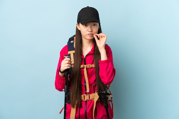 Niña china con mochila y bastones de trekking sobre pared azul aislada mostrando un signo de gesto de silencio