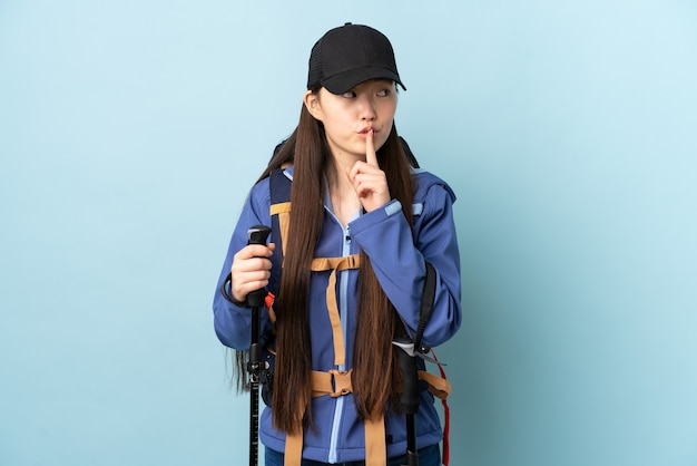 Niña china con mochila y bastones de trekking sobre pared azul aislada que muestra un gesto de silencio poniendo el dedo en la boca