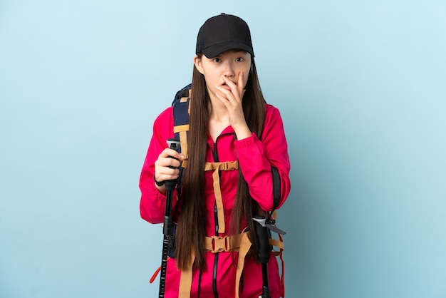 Niña china con mochila y bastones de trekking sobre pared azul aislada sorprendida y conmocionada mientras mira a la derecha