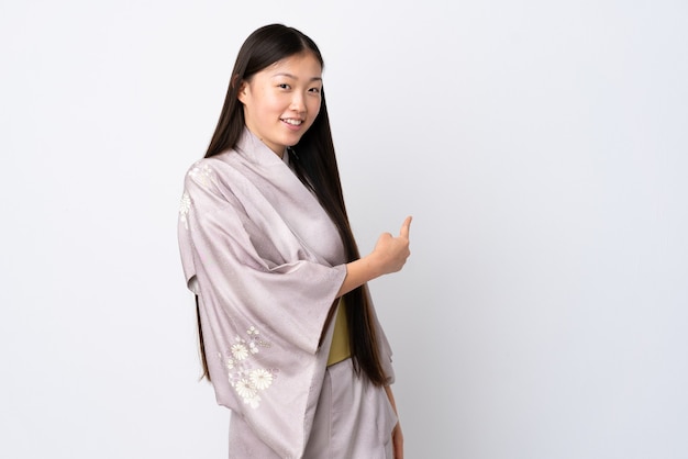 Niña china vistiendo kimono sobre pared aislada apuntando hacia atrás