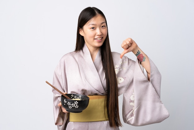 Niña china vistiendo kimono sobre pared blanca aislada orgullosa y satisfecha de sí misma mientras sostiene un plato de fideos con palillos