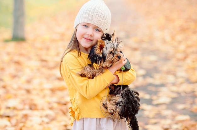 Una niña de cinco años sonríe tiernamente a la cámara en ropa de otoño en octubre y tiene un lindo perrito en sus brazos