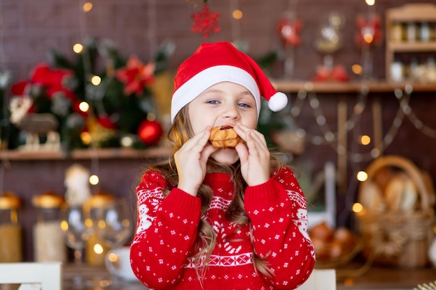 Una niña en la cocina con un árbol de Navidad comiendo galletas de jengibre con un sombrero de Santa Claus, sonriendo y regocijándose preparándose para unas vacaciones, el concepto de Navidad o año nuevo