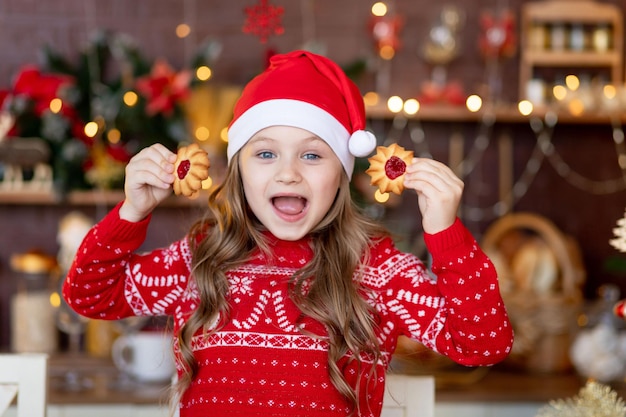 Una niña en la cocina con un árbol de Navidad comiendo galletas de jengibre con un sombrero de Santa Claus, sonriendo y regocijándose preparándose para unas vacaciones, el concepto de Navidad o año nuevo