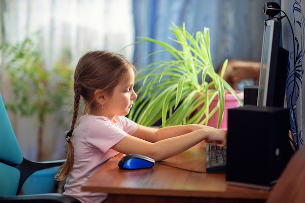 Niña colegiala está sentado en su casa en un escritorio de la computadora y se dedica a una computadora de escritorio.