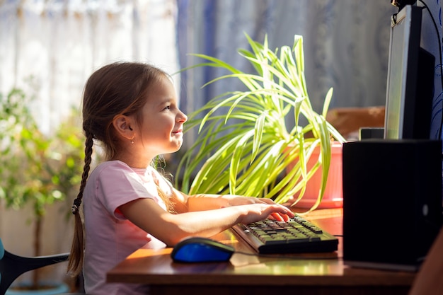 Niña colegiala está sentado en su casa en un escritorio de la computadora y se dedica a una computadora de escritorio.