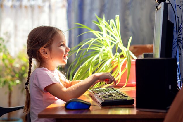 Niña colegiala está sentado en su casa en un escritorio de la computadora y se dedica a una computadora de escritorio.