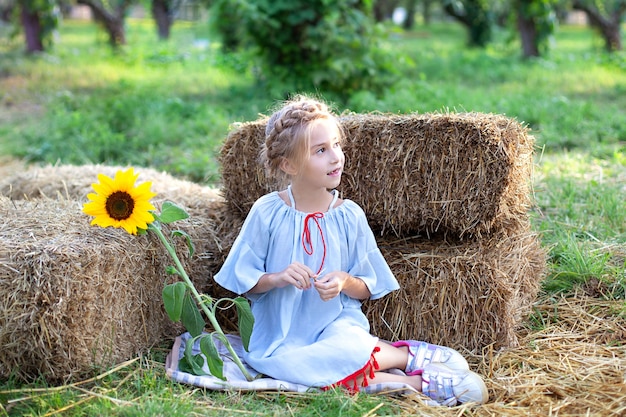 Niña con coleta en la cabeza se sienta en el rollo de pajares en jardín y tiene girasol. Retrato joven rubia con girasol