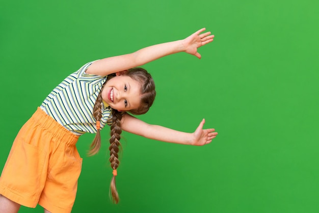 Una niña con coletas mira desde la esquina con los brazos extendidos hacia adelante. Fondo aislado. Copie el espacio. Un niño alegre y alegre sobre un fondo verde. La niña sonríe.