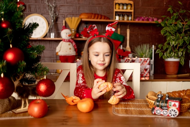 una niña come mandarinas en una cocina oscura junto a un árbol de Navidad con bolas rojas se regocija y sonríe el concepto de año nuevo y Navidad