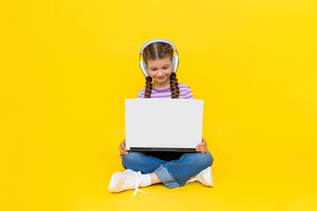 Foto una niña con una computadora portátil una niña pequeña está sentada con las piernas cruzadas en el suelo con los auriculares puestos y sosteniendo una computadora portátil en sus manos redes sociales para adolescentes fondo amarillo aislado