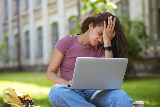 Una niña con una computadora portátil sentada en el césped y con aspecto cansado