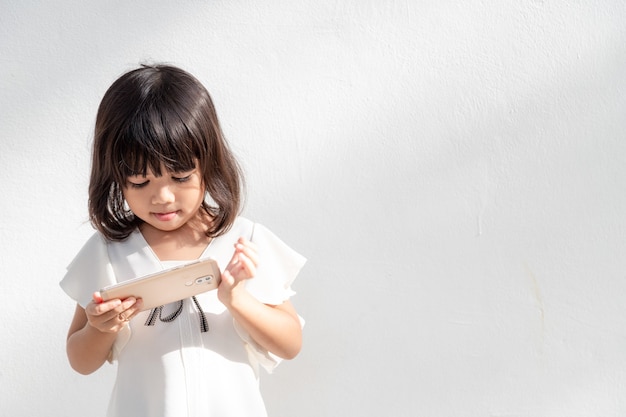 Una niña se concentra en el teléfono, mira el teléfono inteligente, concepto de tecnología para niños, vista de perfil, aislado sobre fondo blanco, espacio de copia