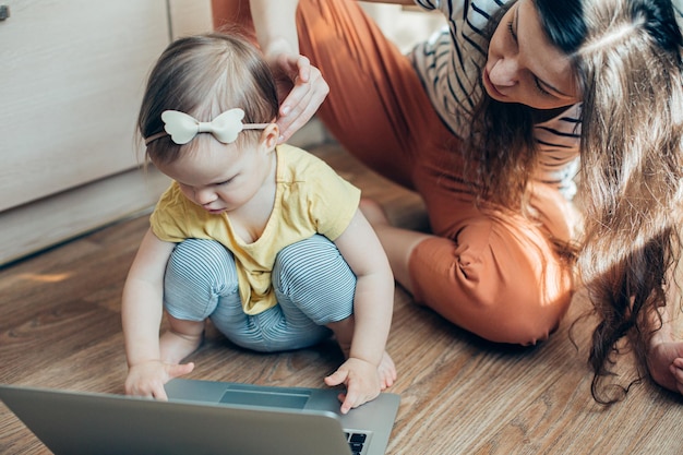 Niña concentrada tocando una laptop moderna en el piso y su madre hablando con ella mientras se sienta cerca