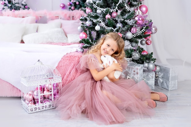 Niña con conejo en el árbol de Navidad con un hermoso vestido rosa, año nuevo y concepto de Navidad