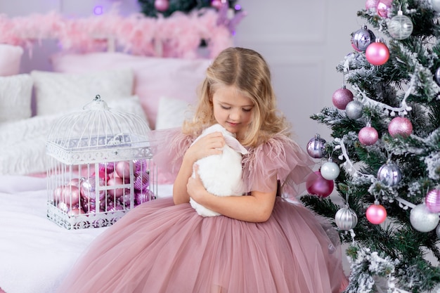 Niña con conejo en el árbol de Navidad con un hermoso vestido rosa