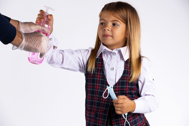 Niña confundida con mascarilla medicinal en la mano y uniforme escolar intenta tomar el desinfectante