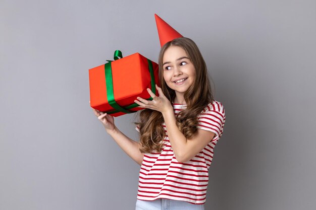 Niña en cono de fiesta sosteniendo una caja de regalo envuelta en azul que está interesada en el interior blanco del regalo