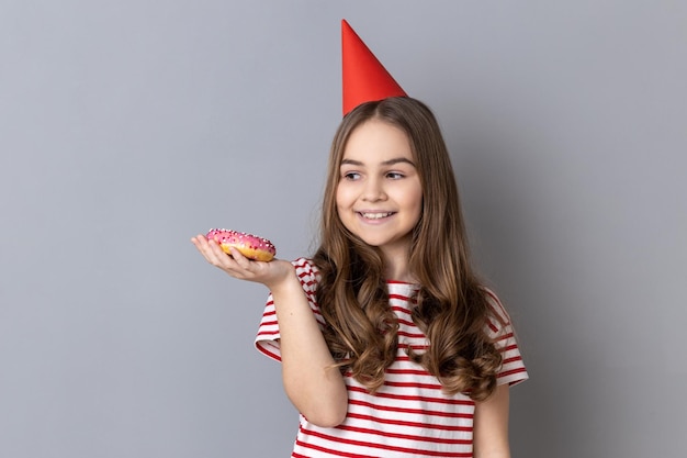 Niña en cono de fiesta sosteniendo en la palma sabroso dulce donut mirando sonriendo al postre