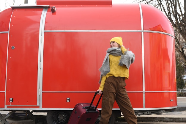 Niña corre con una maleta roja. Gran retro vintage van. Carro viejo. Viajando en el invierno. Chica en un sombrero amarillo brillante y suéter de punto. Concepto de viaje espacio de copia