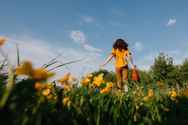 Una niña en un cotte amarillo corre a través de un campo con flores silvestres amarillas en flor con los brazos extendidos ...