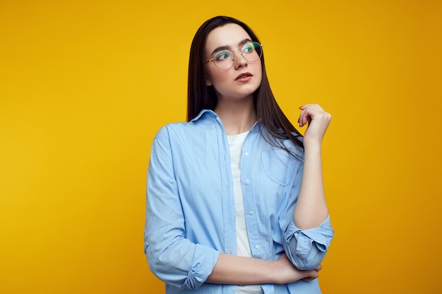 Niña cruza el brazo por encima de la cintura, mira hacia el lado izquierdo, usa camisa azul sobre una pared amarilla