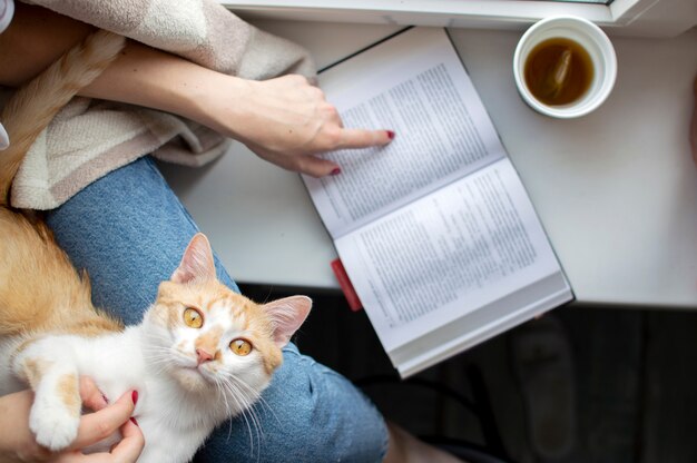 Niña cubierta con una manta se sienta en la ventana con su gato, lee un libro y bebe té, le enseña al gato a leer, copia espacio para el texto