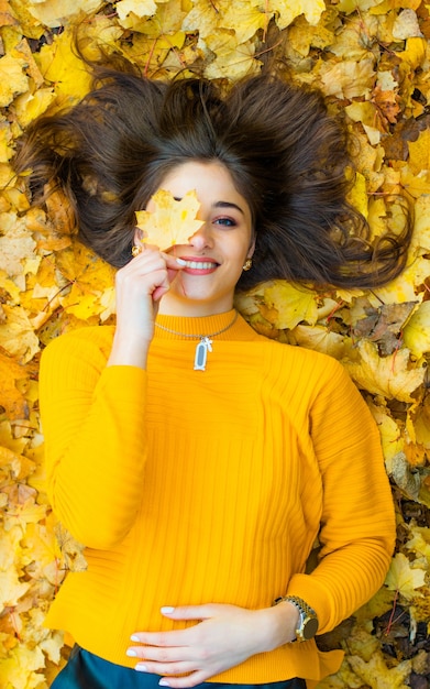 Una niña con un cuello alto amarillo se encuentra en las hojas de otoño cubre sus ojos con una hoja