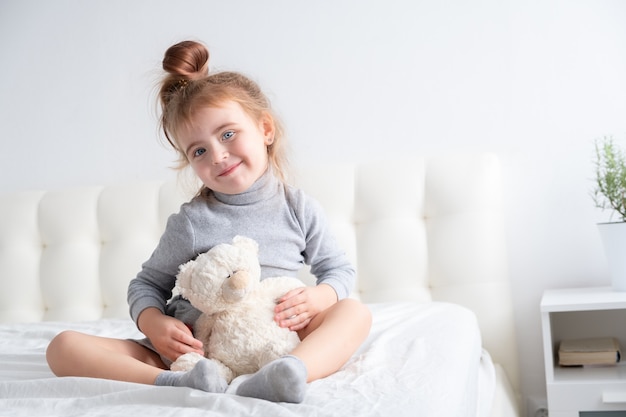 Niña de cuello alto gris jugando con osito de peluche en ropa de cama blanca.