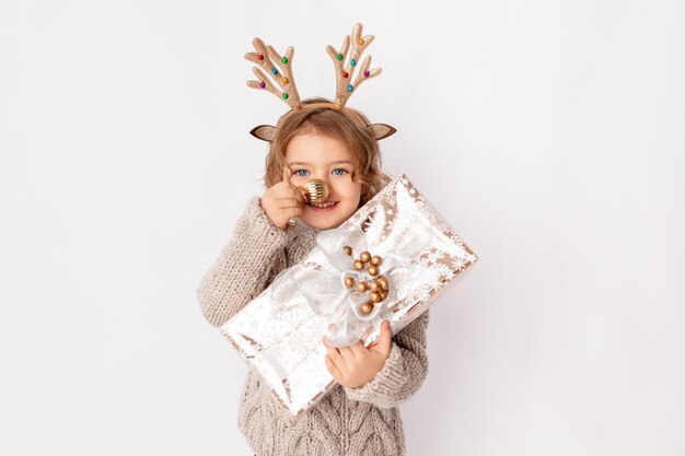 Niña con cuernos de ciervo y bola de Navidad en la nariz sobre un fondo blanco con un regalo en sus manos, espacio para texto, año nuevo y concepto de Navidad