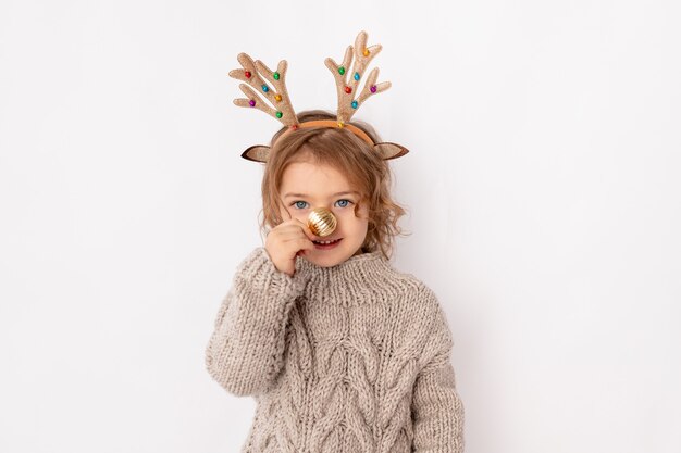 Niña con cuernos de ciervo y bola de Navidad en su nariz sobre fondo blanco, espacio para texto, año nuevo y concepto de Navidad