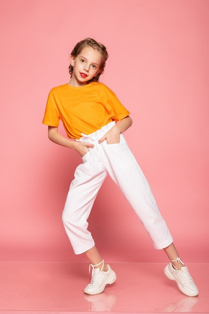 Niña de cuerpo entero en la pared rosada del estudio. vistiendo una camiseta naranja y pantalones blancos y zapatillas blancas.