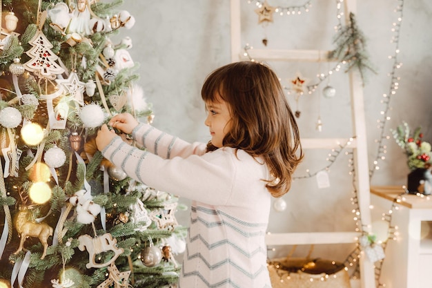 Niña decora el árbol de Navidad con juguetes. Un lindo bebé se está preparando en casa para la celebración de la Navidad.