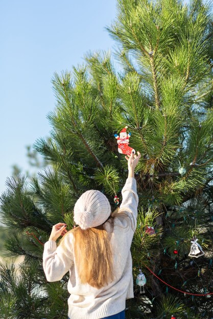 Niña decora con juguetes decorativos y guirnaldas un árbol de año nuevo verde en la calle en invierno en el bosque. Decoraciones para árboles de Navidad.
