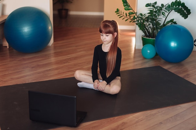 La niña se dedica a la gimnasia y los deportes en línea, un niño se sienta en una alfombra deportiva con una computadora portátil en un