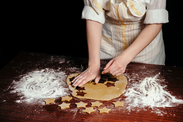 La niña en un delantal corta galletas en forma de masa en forma de estrellas sobre una mesa de madera.