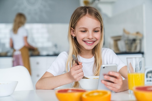 Niña desayunando mientras usa el teléfono en la cocina de casa