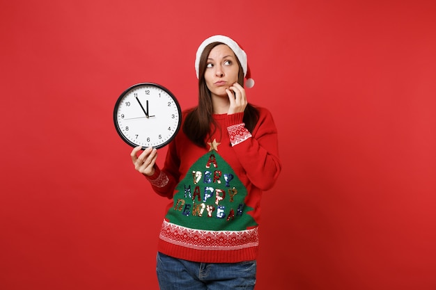 La niña desconcertada de Santa mantenga el reloj redondo, mirando hacia arriba, coloque la mano en la barbilla aislada sobre fondo rojo. Se acaba el tiempo. Feliz año nuevo 2019 celebración concepto de fiesta navideña. Simulacros de espacio de copia.