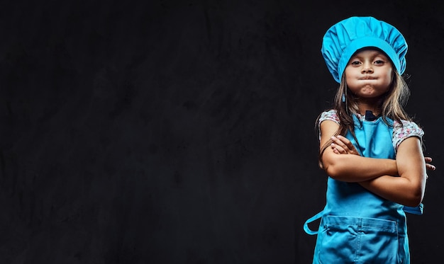 Niña descontenta vestida con uniforme de cocinero azul de pie con los brazos cruzados. Aislado en un fondo de textura oscura.