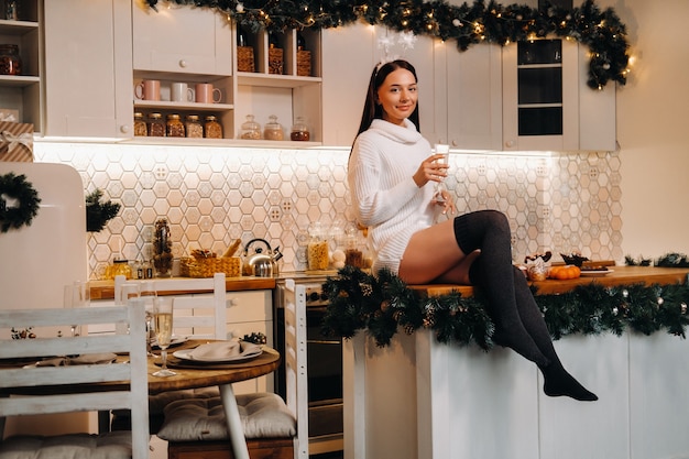 Una niña en el día de Navidad está sentada en la mesa de la cocina y sosteniendo una copa de champán.Mujer en la víspera de año nuevo con champán