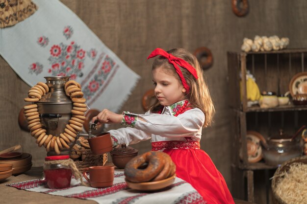 Niña con diadema roja y camisa ornamental vertiendo té de samovar celebrando Maslenitsa
