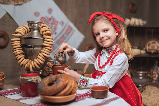 Niña con diadema roja y camisa ornamental vertiendo té de samovar celebrando Maslenitsa