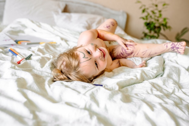 Niña dibuja un arco iris sobre papel blanco con rotuladores en la cama. los niños juegan por la mañana en casa. travieso bebé travieso, manos, pies y cara manchados de pinturas, sucio.