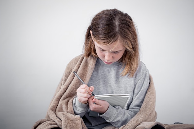 Niña dibuja en un cuaderno envuelto en un plaid sobre un fondo blanco.