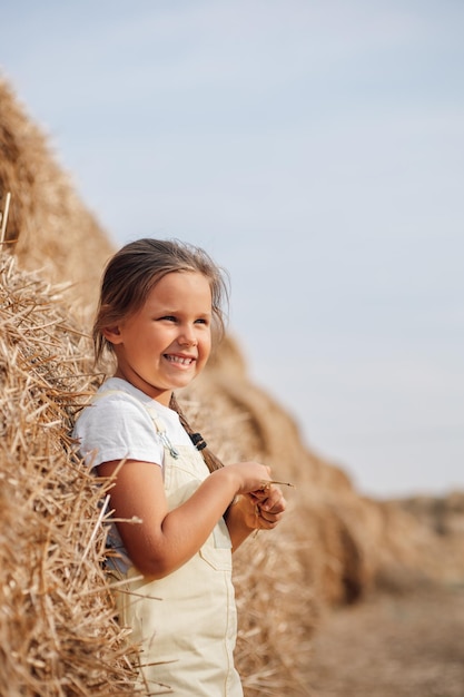 Una niña dichosa parada cerca de un alto heno en el campo con trenzas en las manos de los hombros sosteniendo heno con los ojos entrecerrados mirando a algún lugar lejano Tiempo lejos de la ciudad en el campo