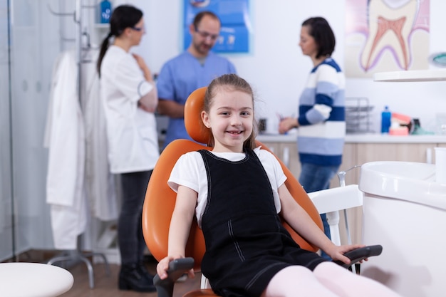 Niña con diente perdido sentado en una silla en la oficina del dentista sonriendo a la cámara. Niño con su madre durante el chequeo de dientes con estomatólogo sentado en una silla.