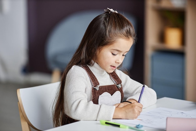 Niña diligente con cabello largo oscuro dibujando con lápices de colores sobre papel mientras se sienta a la mesa en cla